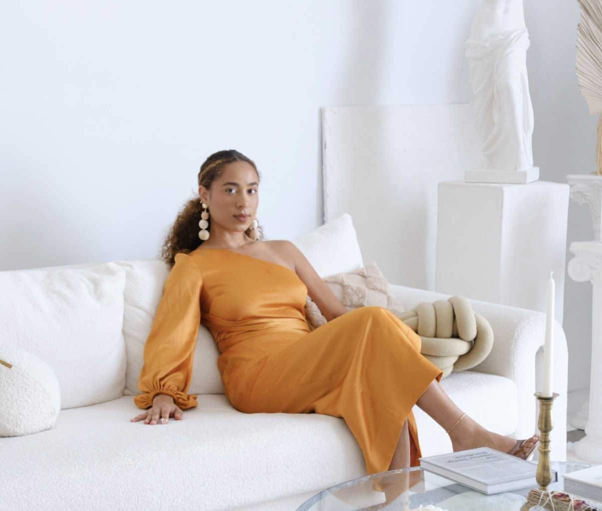 A woman in a one-shoulder mustard-colored dress sits on a white sofa in a bright, minimalist room with white décor and sculptures.